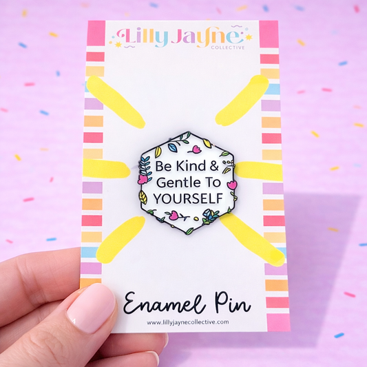 Enamel pin with 'Be Kind & Gentle To YOURSELF' message on a colorful card, held by a hand against a pink background.