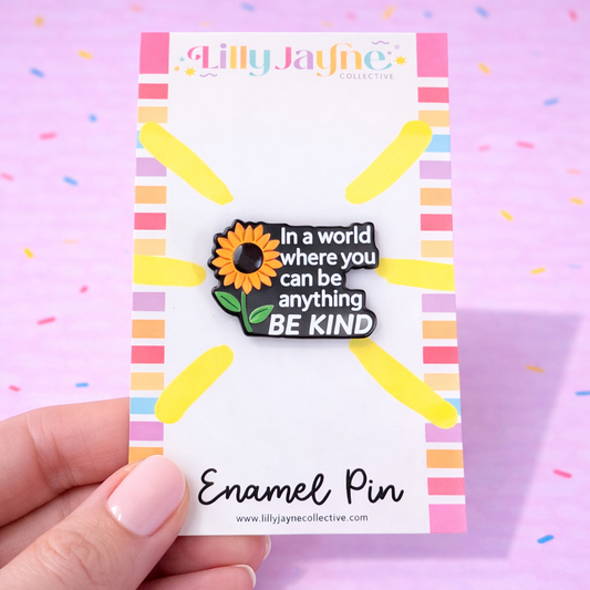 Enamel pin with sunflower design and text on a colorful card, held by a hand against a pink background.