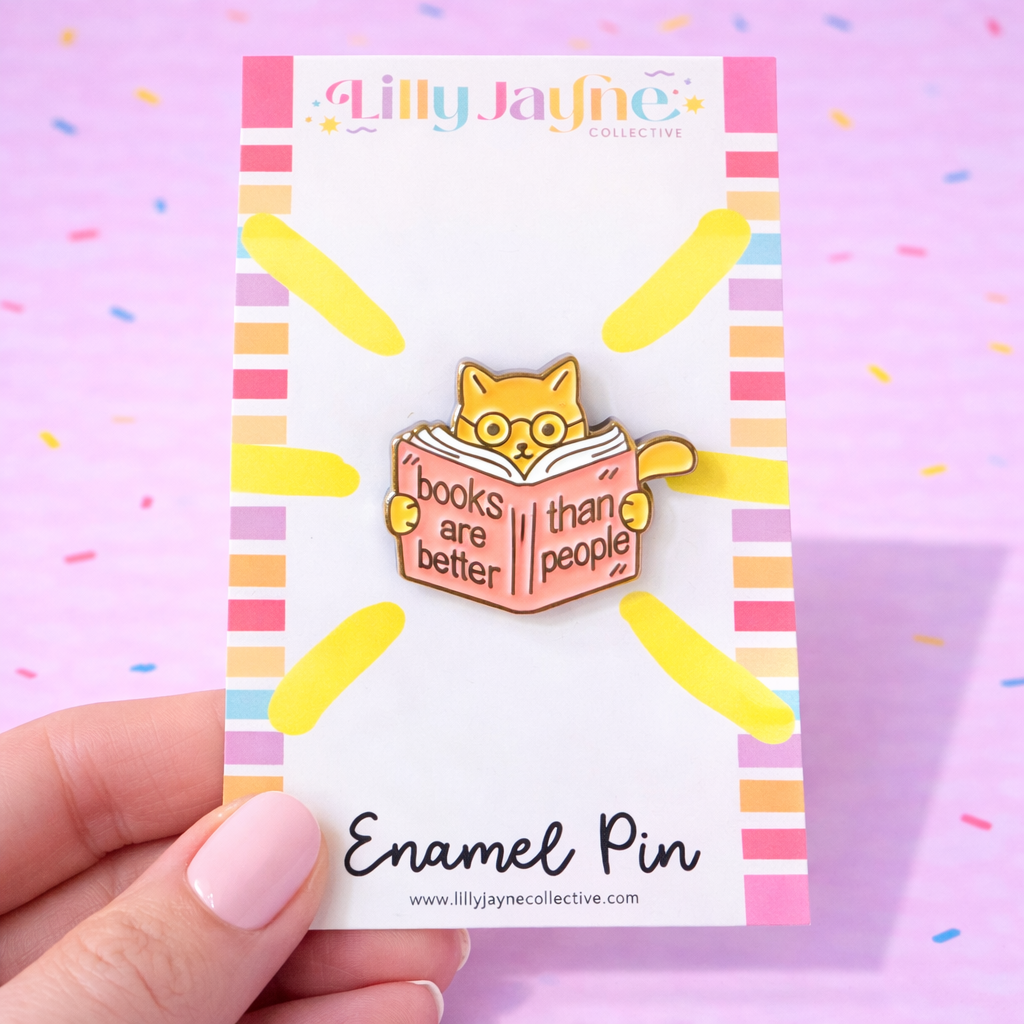 Enamel pin with a cat reading a book design on a colorful card, held by a hand against a pink background.
