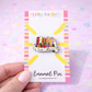 Enamel pin on a colorful card held by a hand with a pastel heart pattern background