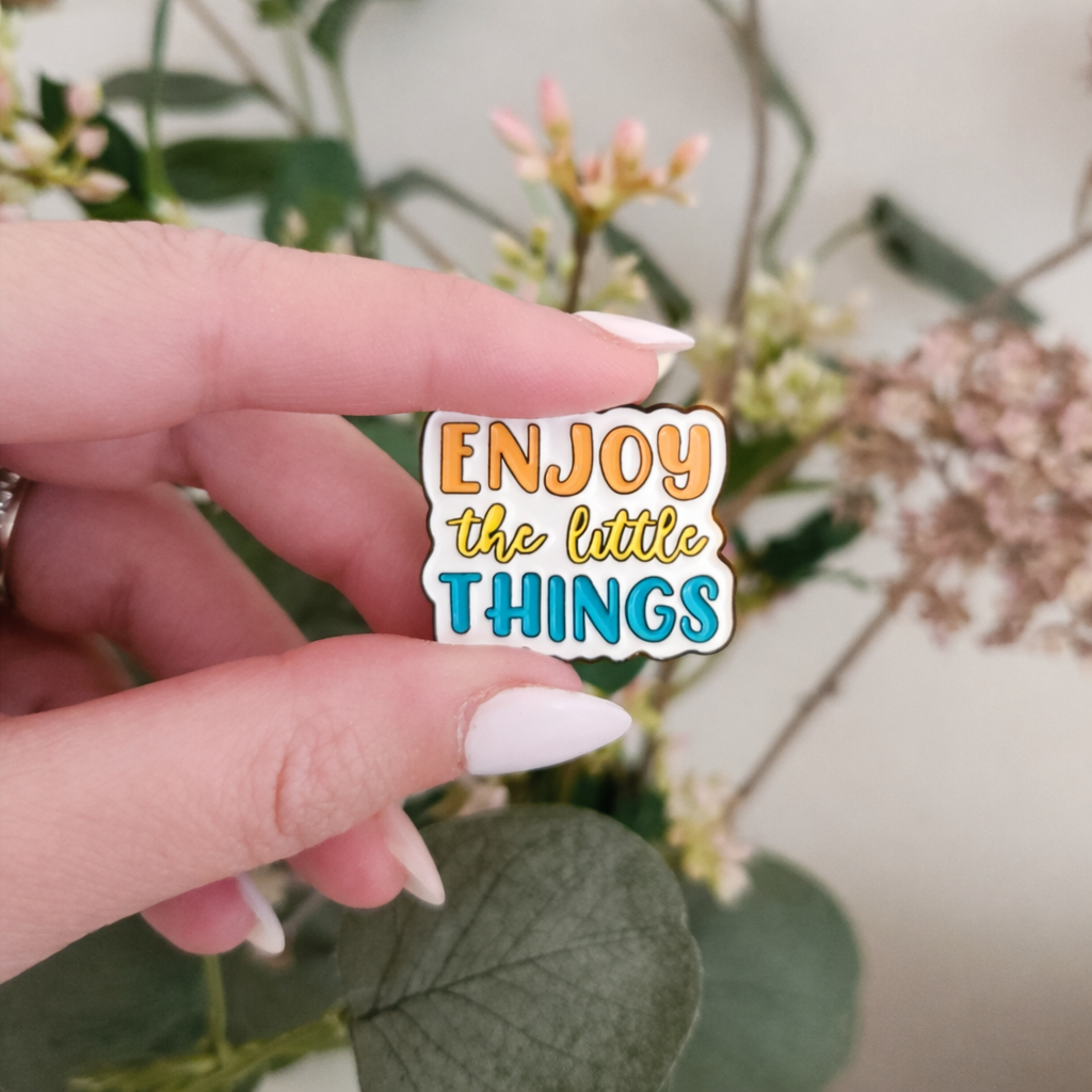 Hand holding a 'Enjoy the little things' pin with a floral background