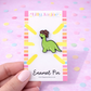 Lilly Jayne enamel pin with a green dinosaur design on a colorful card against a pink background with polka dots.