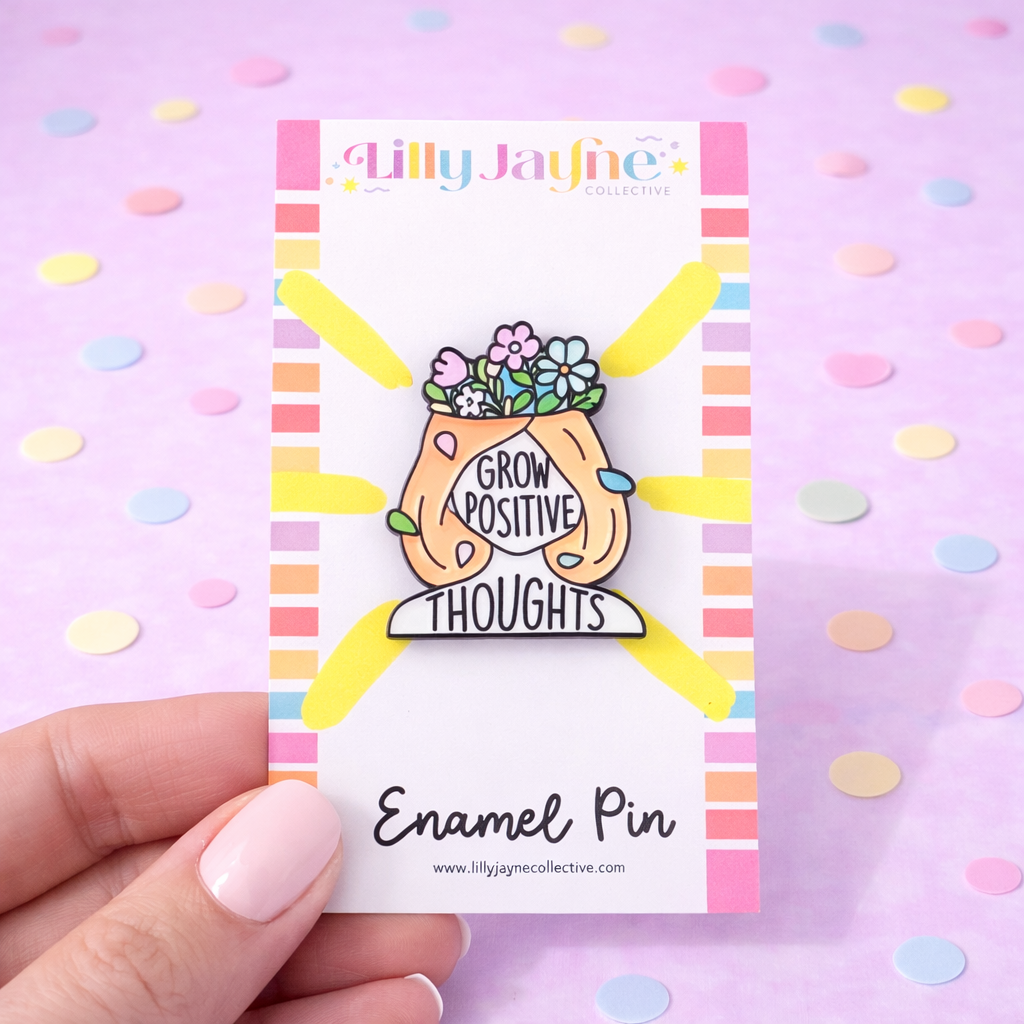Enamel pin with 'Grow Positive Thoughts' design on a colorful card, held against a pink background with polka dots.