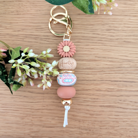 Handmade keyring with deep pink daisy flower charm, engraved 'Aunty' wooden bead, floral patterned bead, and pastel pink accents on a wooden surface with greenery
