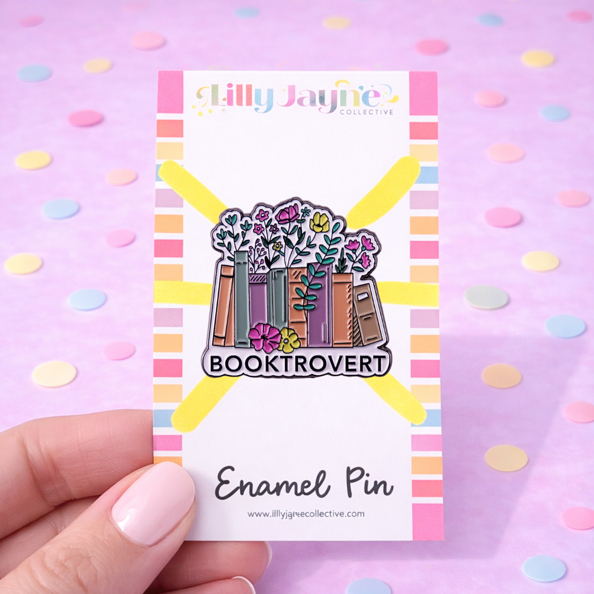 Enamel pin with 'Booktrovert' design on a colorful card, held against a pink background with polka dots.