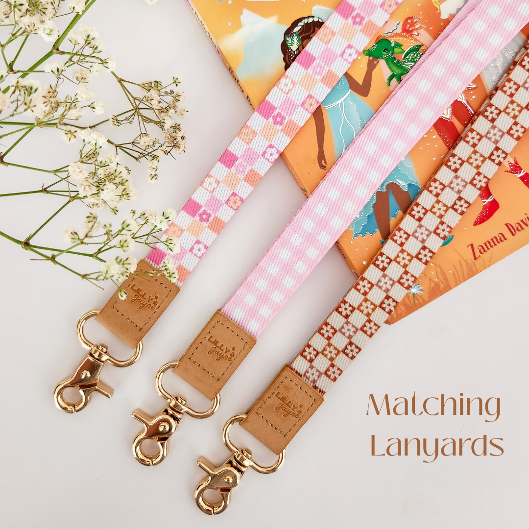 Three colourful fabric Lanyards with gold hardware and tan leather ends, featuring pink gingham daisy, blush polka dot stripe, and retro orange checker floral patterns, laid on pastel notebooks with floral accents – Lilly Jayne Collective.