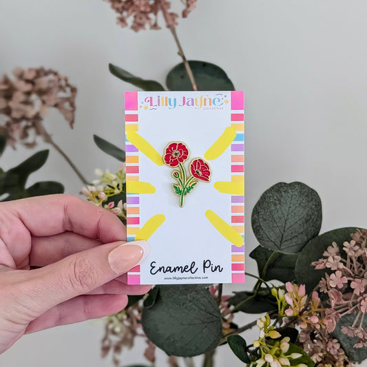 Hand holding a card with a floral enamel pin and 'Lilly Jayne' branding, surrounded by flowers and leaves.