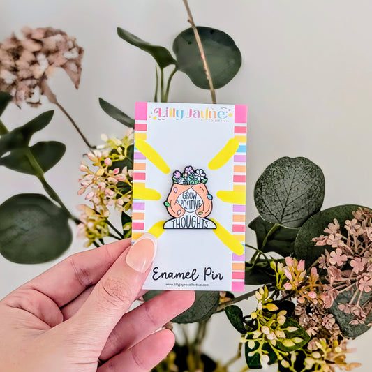 Hand holding a card with an enamel pin featuring a plant and text, surrounded by flowers and leaves.