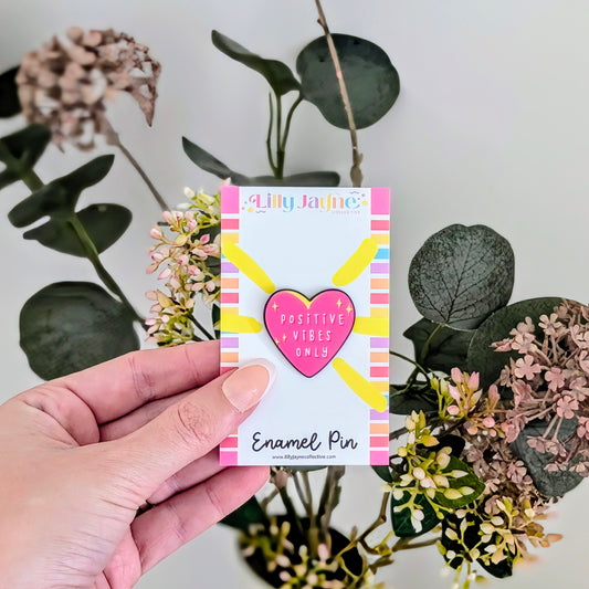 Hand holding a card with an enamel pin featuring a heart and text, set against a background of greenery.