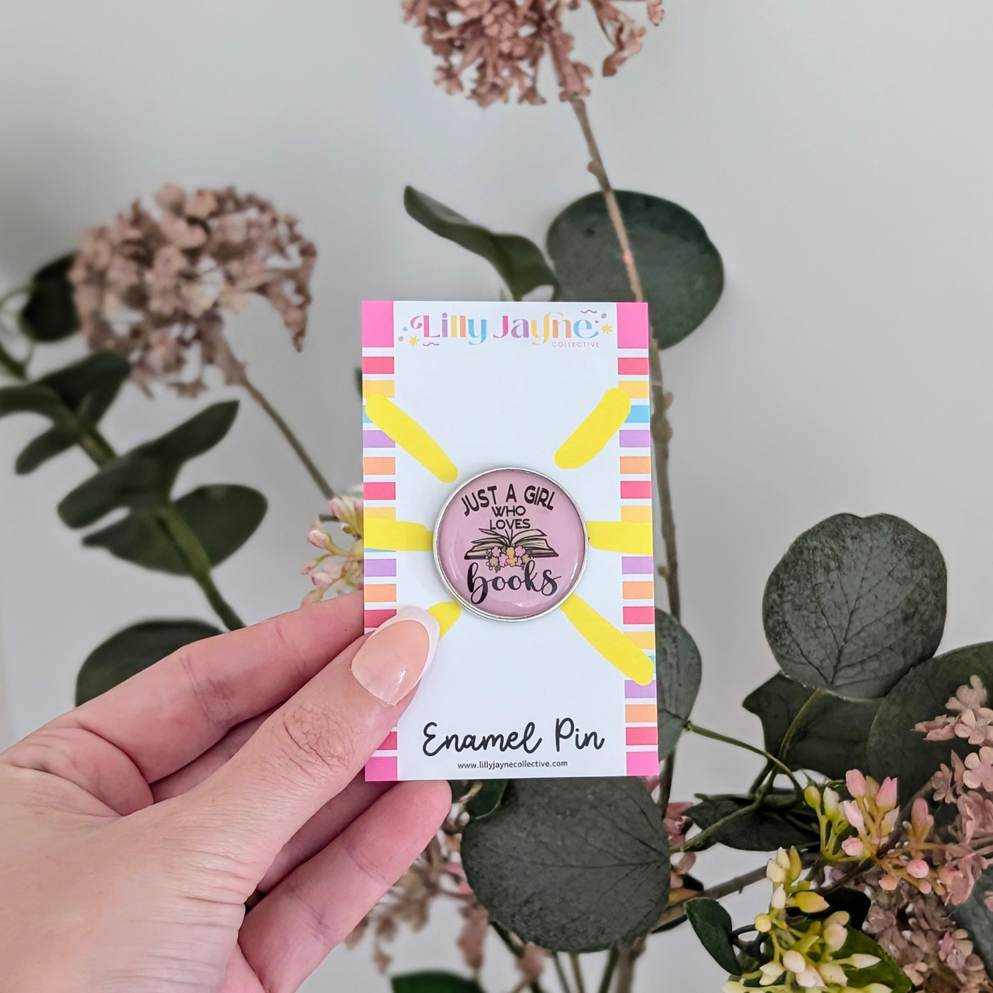 Hand holding a card with an enamel pin featuring a sun design and text, against a floral background.