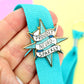 Enamel pin with 'Proudly Neuro-Sparkly' text on a blue ribbon held by a hand against a colorful background.
