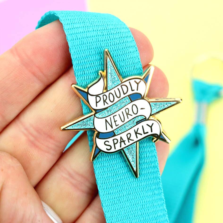 Enamel pin with 'Proudly Neuro-Sparkly' text on a blue ribbon held by a hand against a colorful background.
