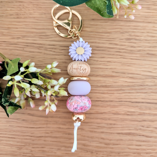 Handmade keyring with purple daisy flower charm, engraved 'Aunty' wooden bead, floral patterned bead, and pastel pink accents on a wooden surface with greenery