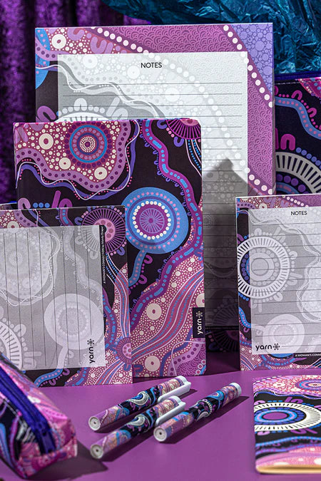 Colorful stationery items including notebooks and pens on a purple - aboriginal art surface