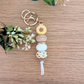 Handmade keyring with yellow sunflower flower charm, engraved 'Aunty' wooden bead, floral patterned bead, and pastel pink accents on a wooden surface with greenery