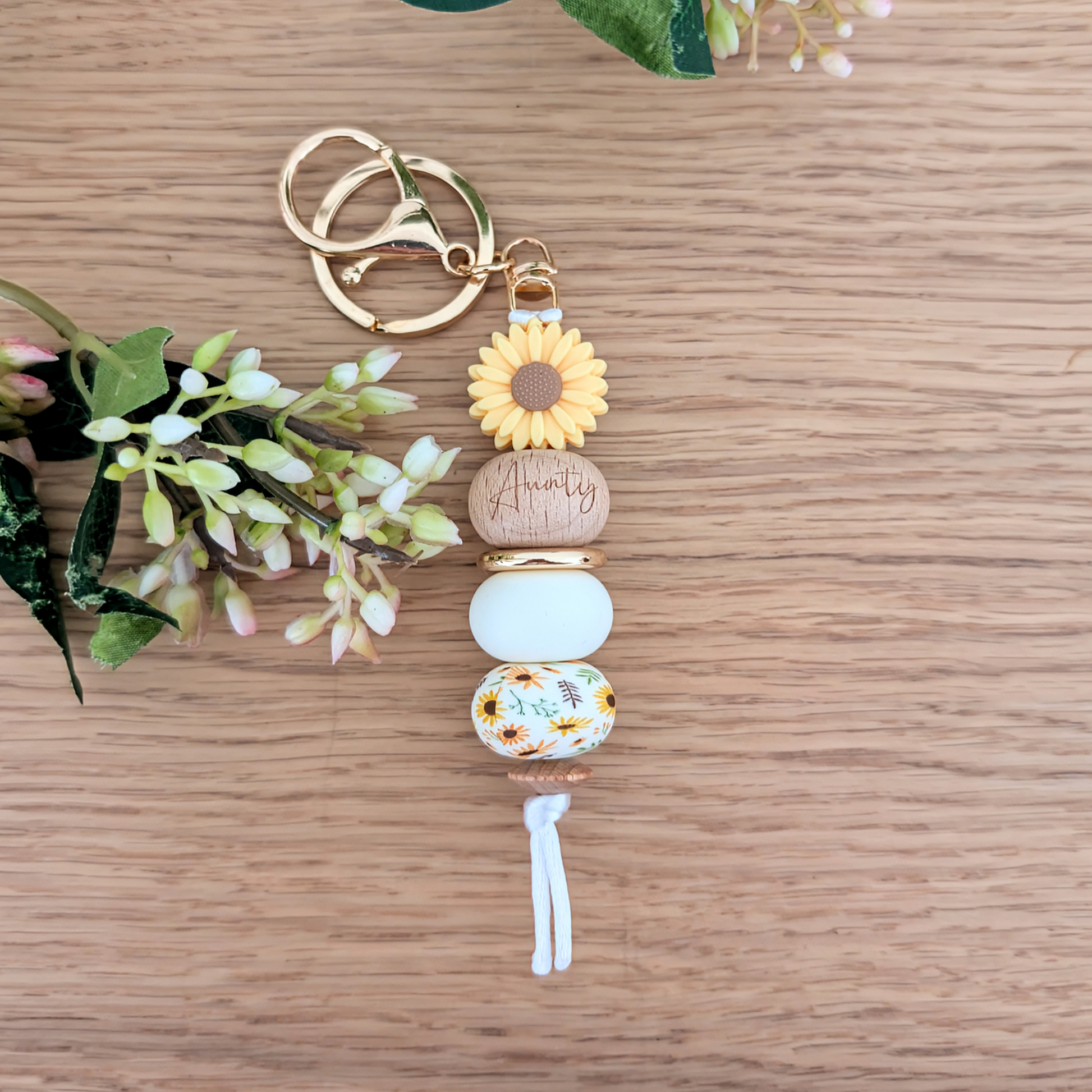 Handmade keyring with yellow sunflower flower charm, engraved 'Aunty' wooden bead, floral patterned bead, and pastel pink accents on a wooden surface with greenery