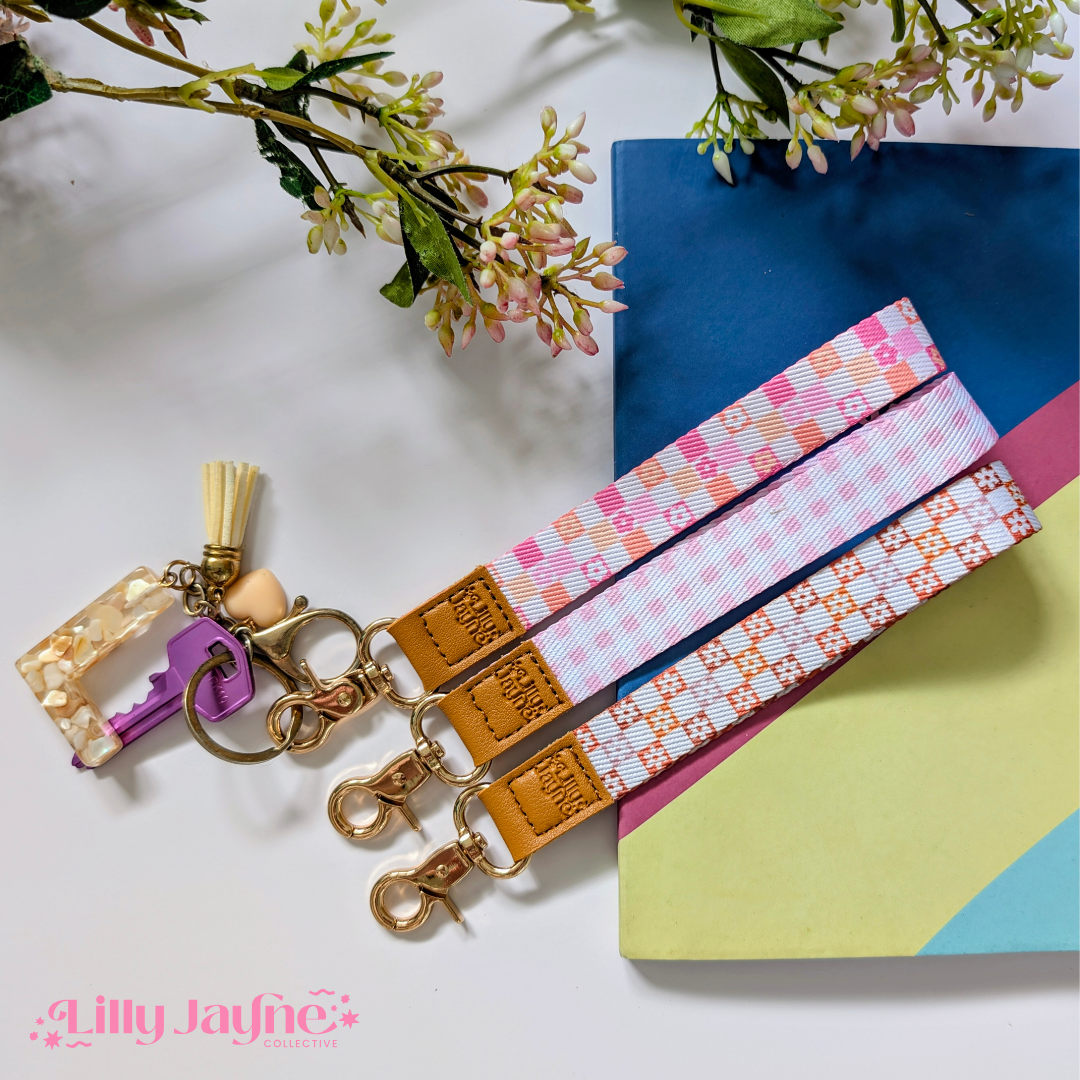 Three colourful fabric wristlet keychains with gold hardware and tan leather ends, featuring pink gingham daisy, blush polka dot stripe, and retro orange checker floral patterns, laid on pastel notebooks with floral accents – Lilly Jayne Collective.