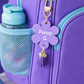 Purple backpack with a water bottle and a personalized purple flower charm on a light background