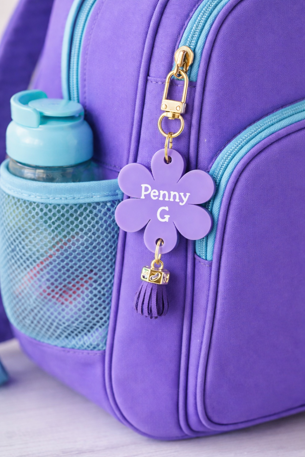 Purple backpack with a water bottle and a personalized purple flower charm on a light background