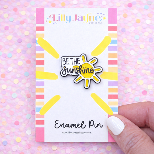 Enamel pin with 'Be the Sunshine' design on a colorful card, held against a polka dot background.