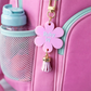 Pink backpack with a flower-shaped charm labeled 'Ruby W' and a water bottle.