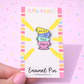 Enamel pin with 'We Are All Mad Here' quote on a colorful card, held against a pink background.