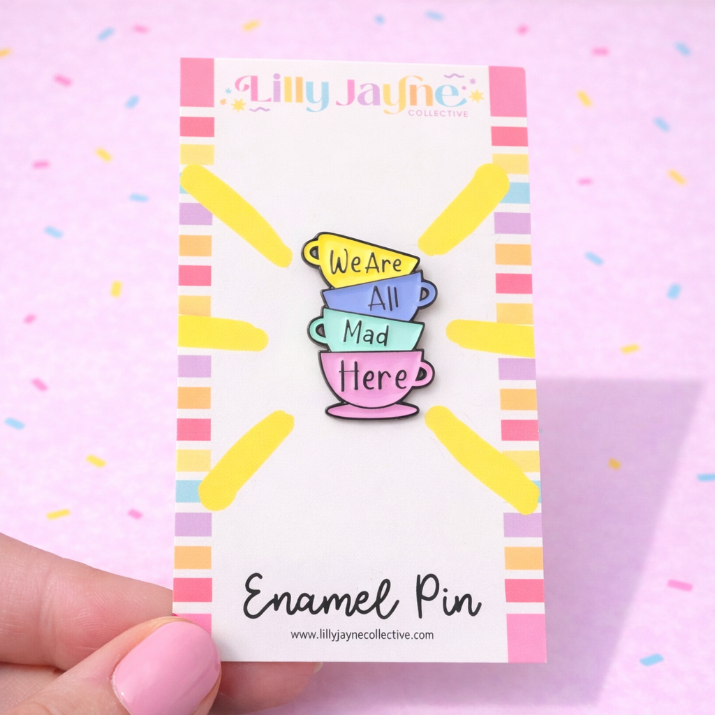 Enamel pin with 'We Are All Mad Here' quote on a colorful card, held against a pink background.