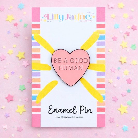 Heart-shaped enamel pin with 'Be a Good Human' text on a colorful card, surrounded by stars on a pink background.