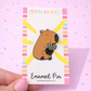 Enamel pin on a card with colorful border, held against a pink background.