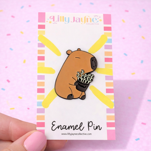 Enamel pin on a card with colorful border, held against a pink background.