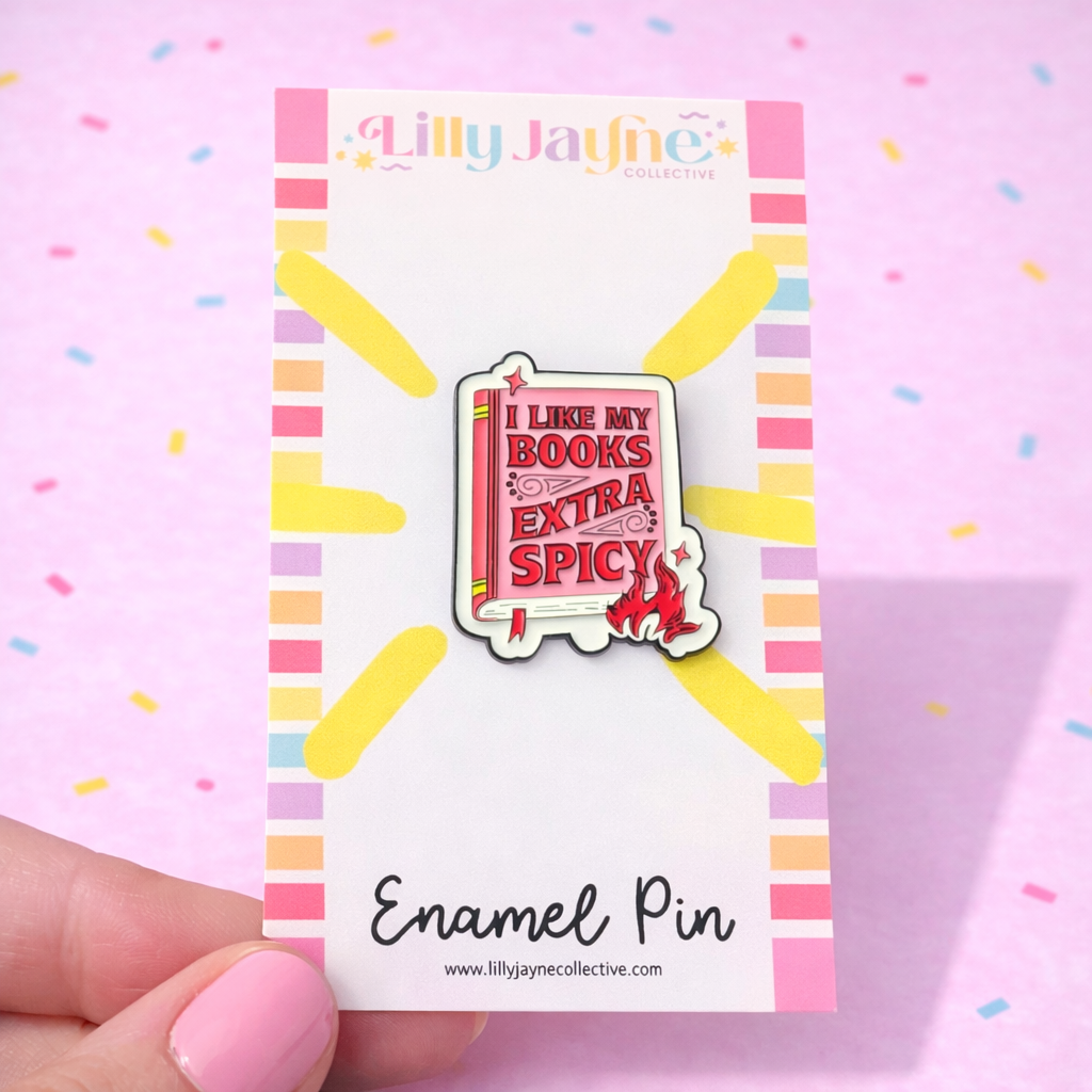Enamel pin with 'I like my books extra spicy' text on a colorful card, held by a hand against a pink background.