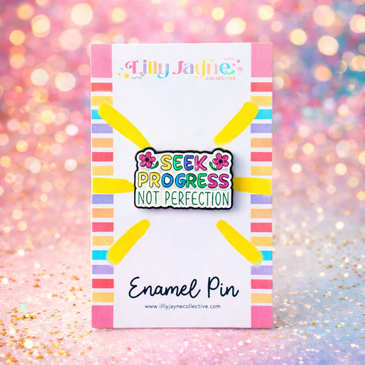Enamel pin with 'Seek Progress Not Perfection' message on a colorful card against a glittery background