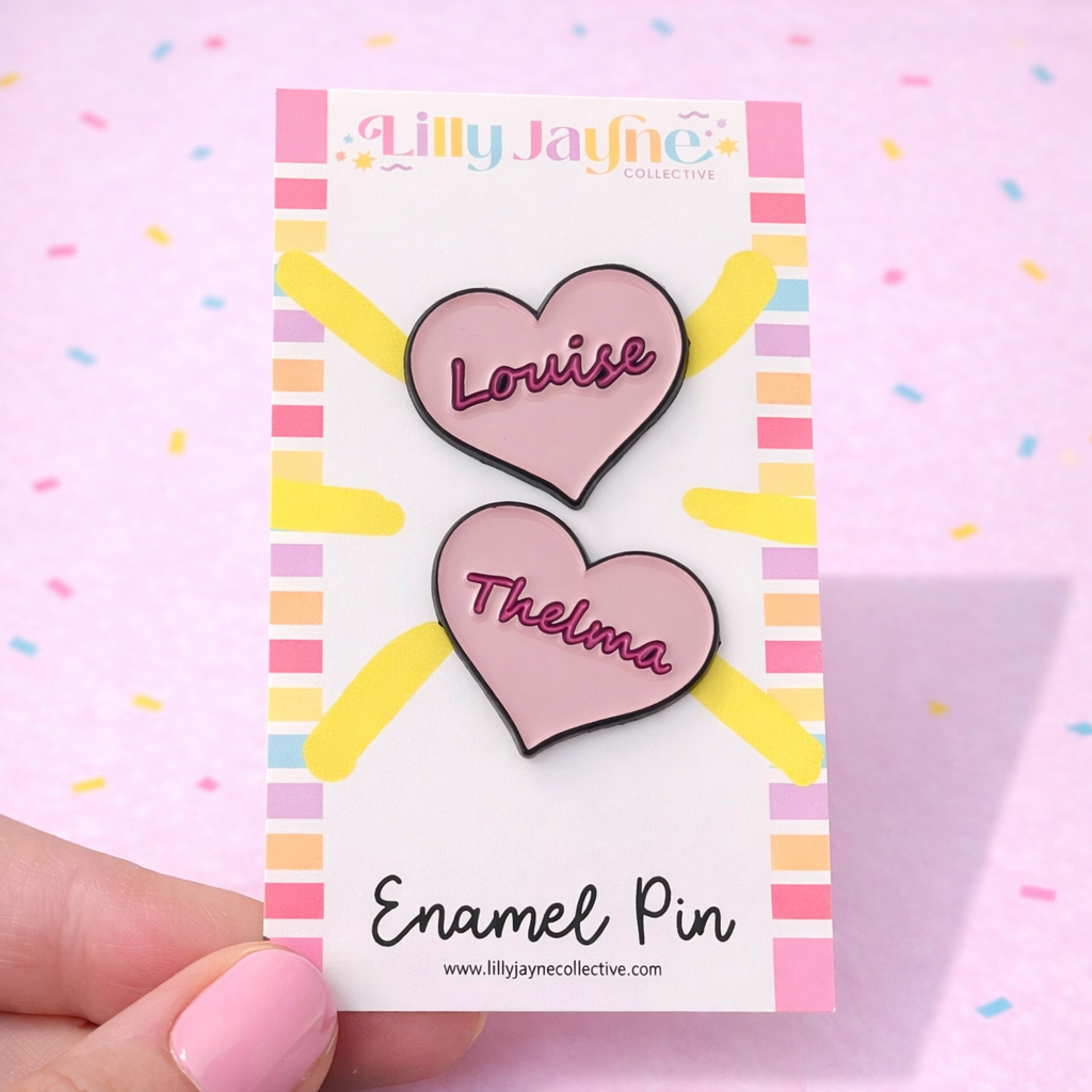 Two heart-shaped enamel pins on a card with 'Lilly Jayne Collective' branding, held against a pink background.