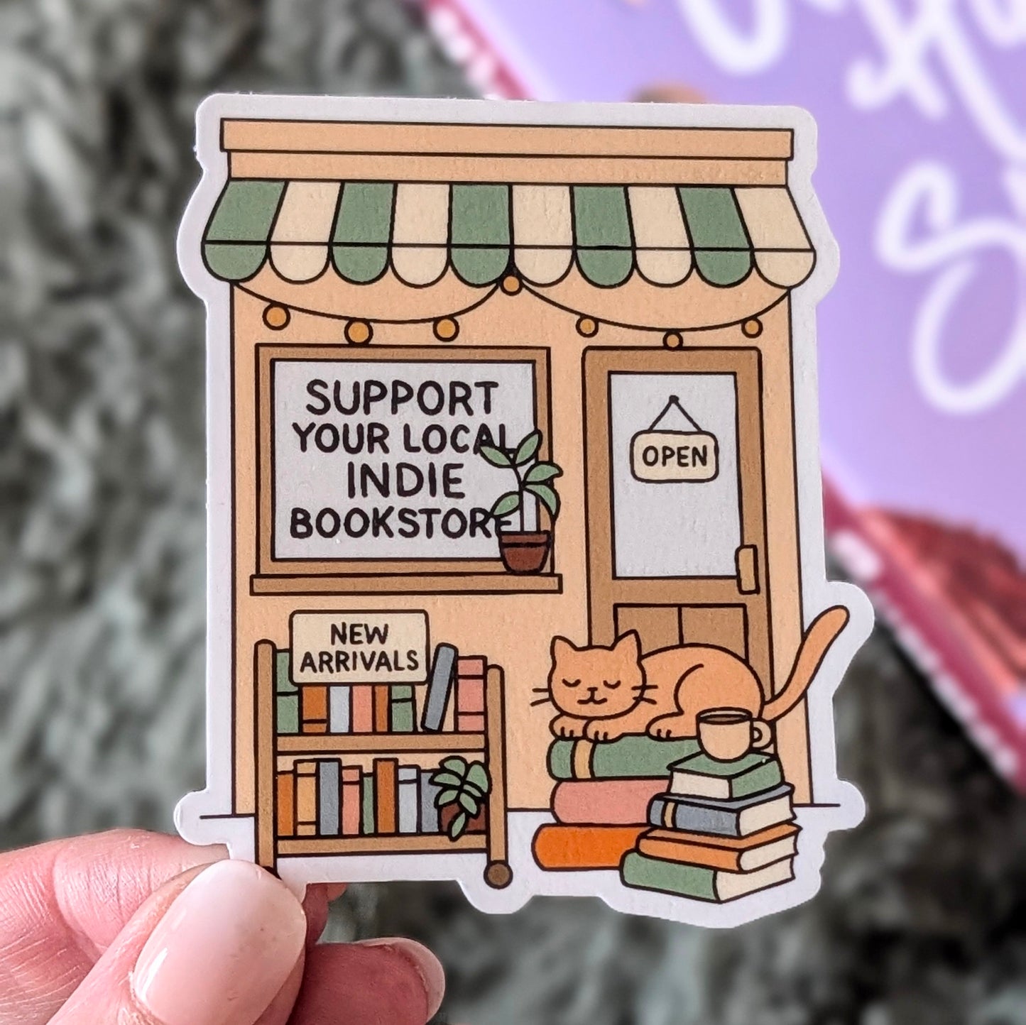Sticker of a local indie bookstore with a cat and books, held by a hand.