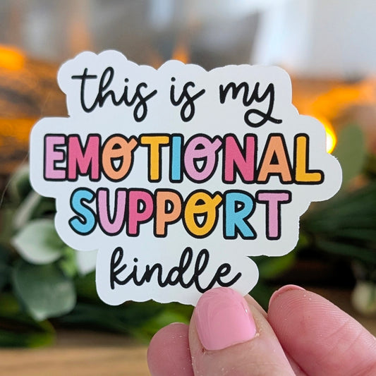 Sticker with colorful text 'this is my emotional support kindle' held by a hand.