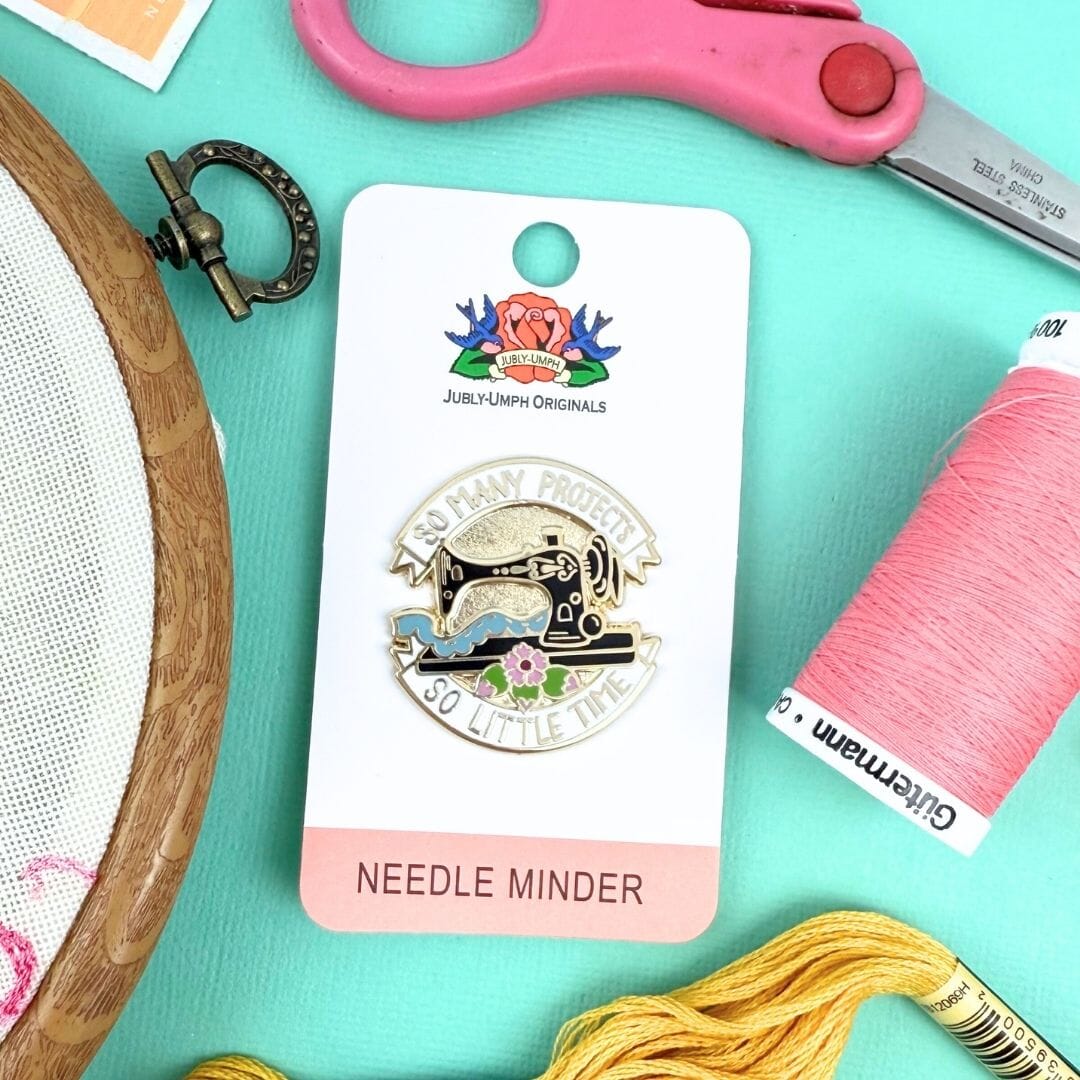 Needle minder on a card with embroidery hoop, scissors, and thread on a turquoise background