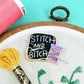 Embroidery hoop with 'Stitch and Bitch' patch, pink thread, and green embroidery on a teal background.