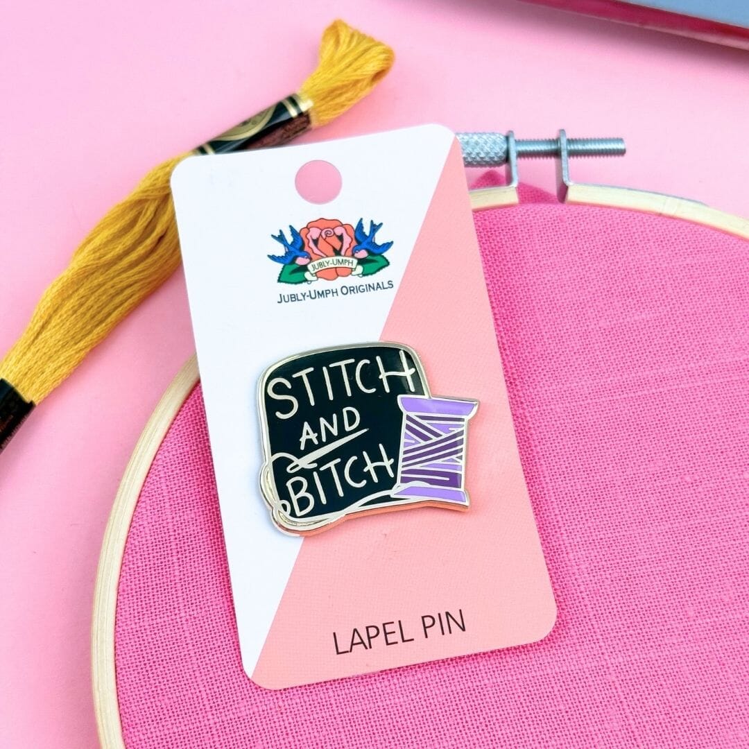 Lapel pin with 'Stitch and Bitch' design on a card with embroidery hoop and thread in the background.